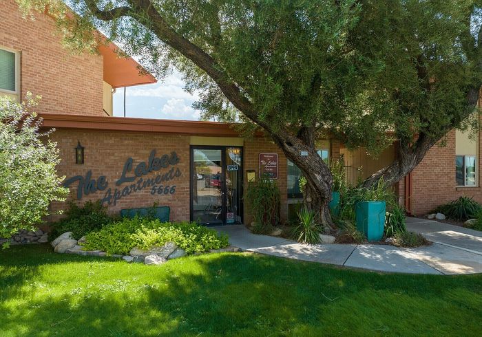 Charming entrance to The Lakes Apartments, with lush greenery and mature trees enhancing the inviting brick facade.