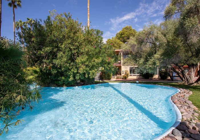 Inviting residential scene with a sparkling swimming pool surrounded by lush greenery and a charming apartment building under a clear blue sky.