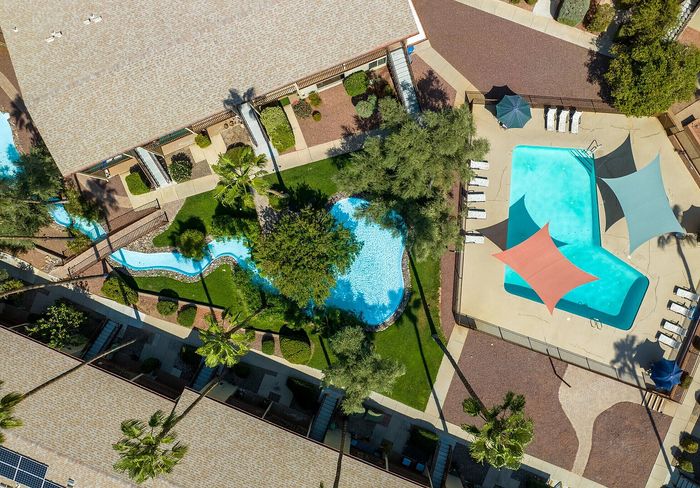 Aerial view of a luxurious apartment complex featuring a large swimming pool with colorful sunshades, lush landscaped gardens, and winding pathways.