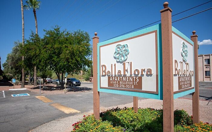 Inviting Bella Flora Apartments sign positioned amidst lush greenery and a sunny, blue sky backdrop.