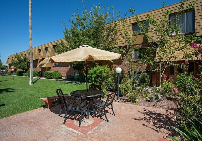 Charming apartment exterior with lush landscaping, brick facade, and inviting patio seating under a large umbrella.