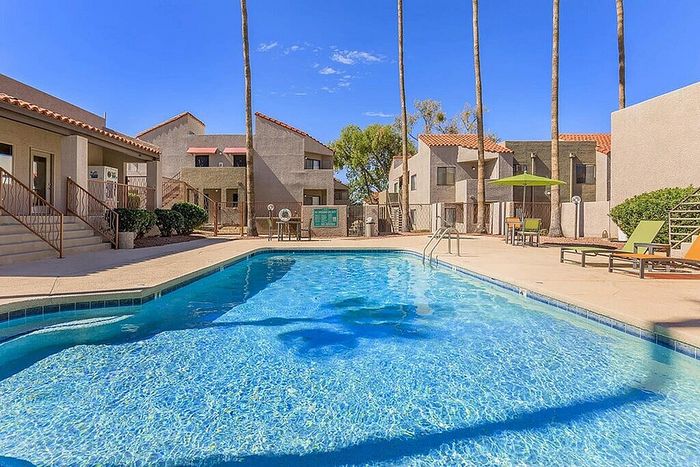 Inviting apartment complex with a crystal-clear pool, surrounded by loungers and lush greenery, under a bright blue sky.