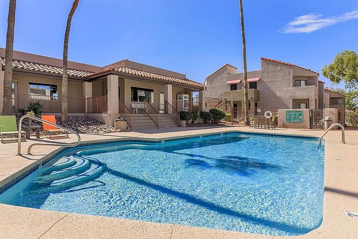 Inviting apartment complex pool area with modern architecture, palm trees, and sun loungers under a clear blue sky.
