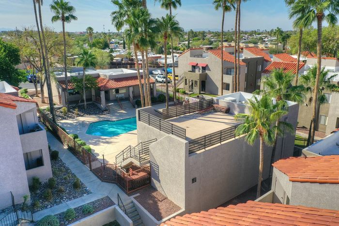 Charming apartment complex featuring a sunlit swimming pool, lush palm trees, and terracotta rooftops, offering a serene resort-style living experience.