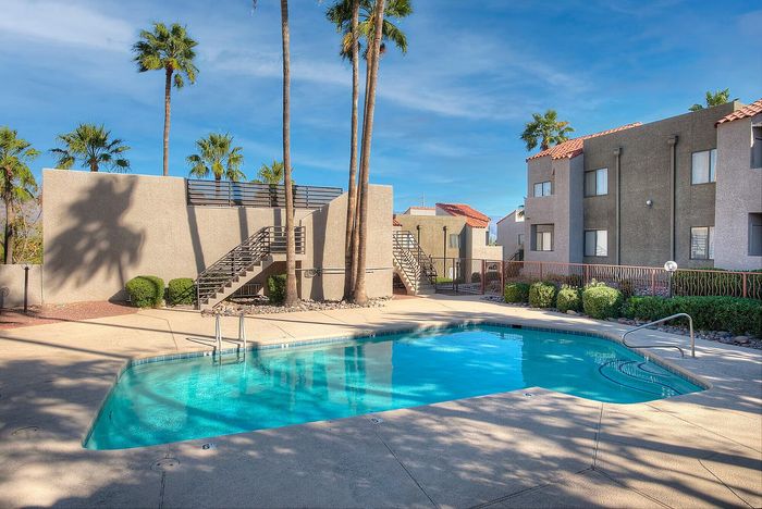 Charming apartment complex with a sparkling outdoor pool, surrounded by lush palm trees and modern architecture under a clear blue sky.