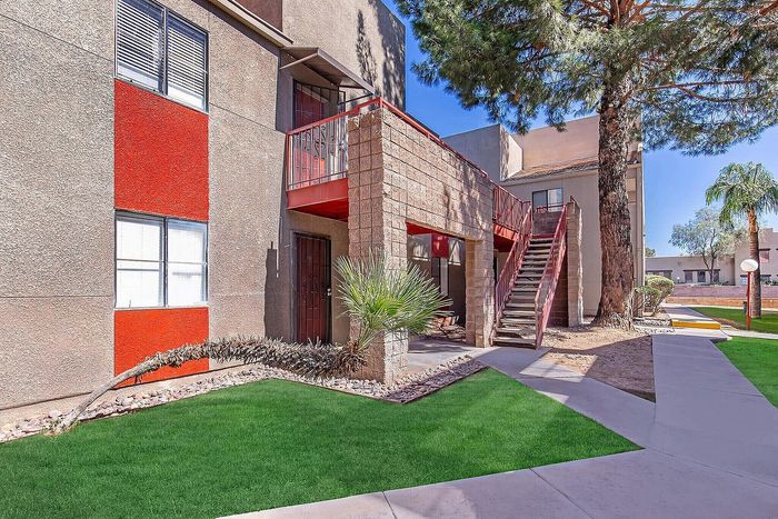 Charming apartment exterior featuring vibrant red accents, lush green landscaping, and a welcoming stairway under tall trees.