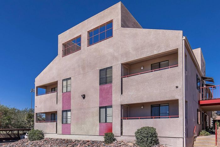 Modern apartment building exterior with unique geometric architecture, red accents, and ample windows against a clear blue sky.