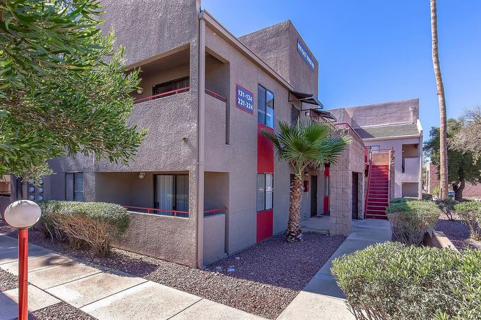 Modern apartment building with vibrant red accents, surrounded by lush greenery and a clear blue sky, highlighting a welcoming entrance.