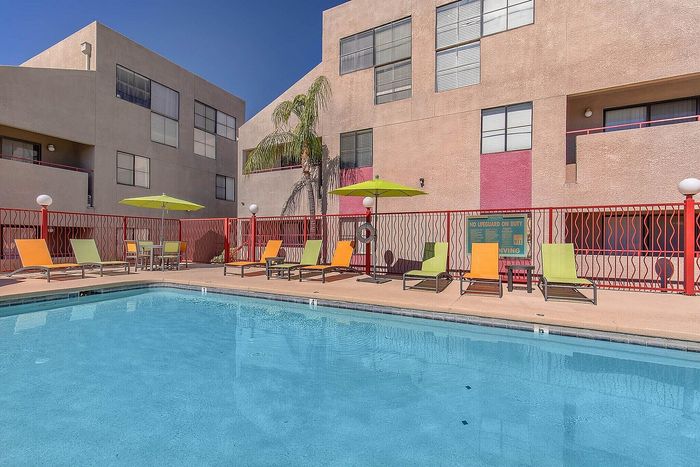 Vibrant apartment complex featuring a sparkling outdoor pool, colorful lounge chairs, and lush palm trees under a clear blue sky.