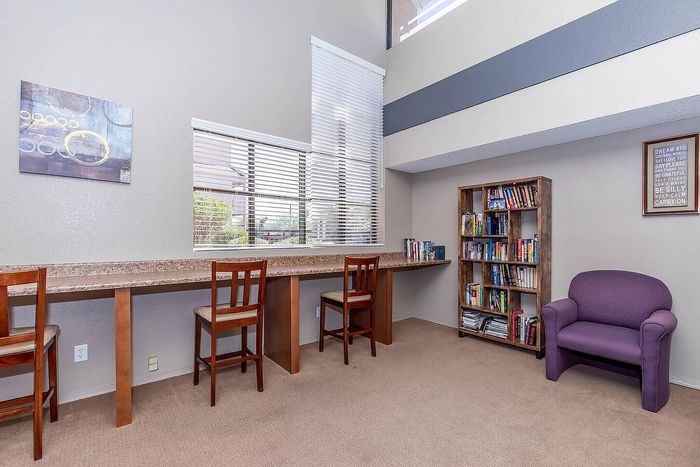 Inviting library nook with a long work desk, three chairs, a cozy purple armchair, and a bookshelf filled with books, bathed in natural light from a generous window.