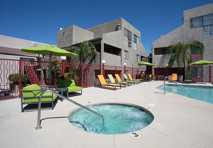 Modern apartment complex with a stunning outdoor pool and spa area, featuring vibrant green and orange lounge chairs and umbrellas, surrounded by palm trees and contemporary architecture.