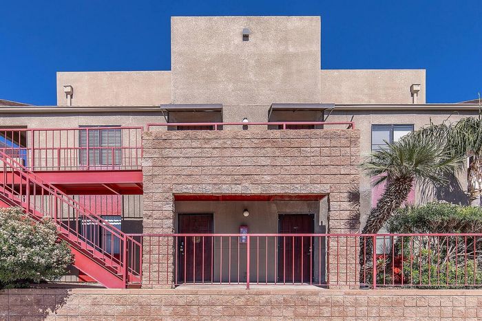 Modern apartment building exterior with bold red accents, lush greenery, and vibrant desert landscaping under a clear blue sky.