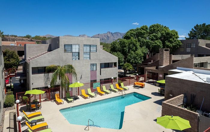 Vibrant apartment complex with a refreshing pool, surrounded by colorful lounge chairs and lush greenery, set against a stunning mountain backdrop.