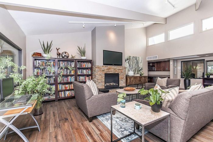 Elegant living room with high ceilings, cozy stone fireplace, and abundant natural light; features chic gray sofas, vibrant indoor plants, and a wall lined with bookshelves.