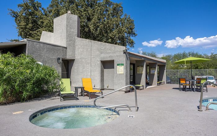 Charming outdoor area featuring a cozy hot tub, modern patio furniture, and lush greenery under a bright blue sky.