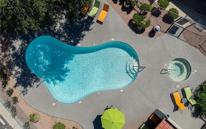 Aerial view of a luxurious apartment pool area with a sparkling kidney-shaped pool, surrounded by colorful lounge chairs and lush greenery.