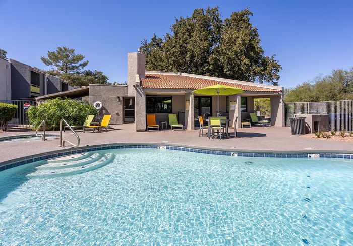 Inviting outdoor pool area with sun loungers and umbrella, surrounded by lush greenery and a modern clubhouse, perfect for relaxation and gatherings.