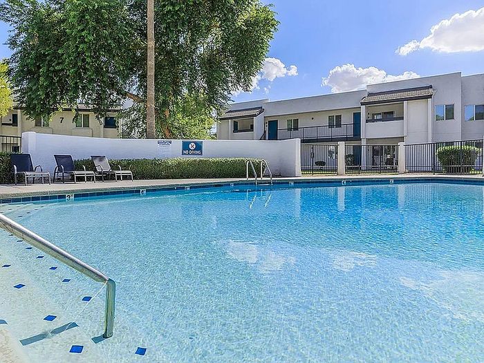Spacious swimming pool surrounded by comfortable lounge chairs and modern apartment buildings under a clear blue sky.