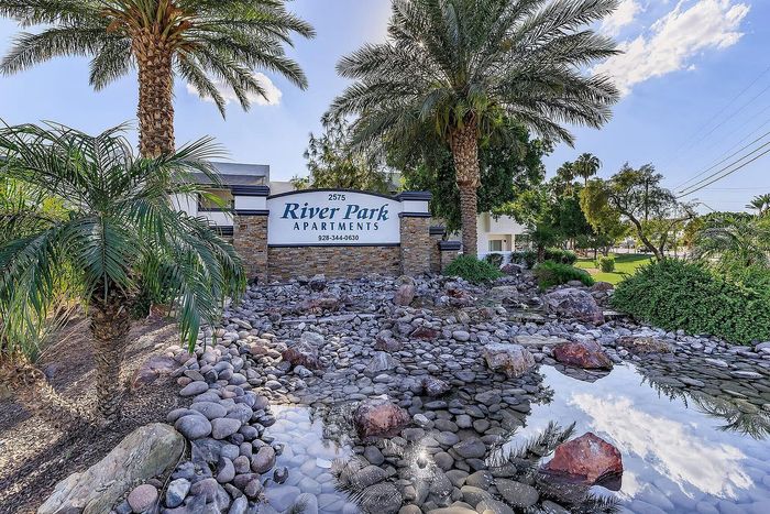 Welcoming entrance of River Park Apartments surrounded by lush greenery and serene stone-lined water features.
