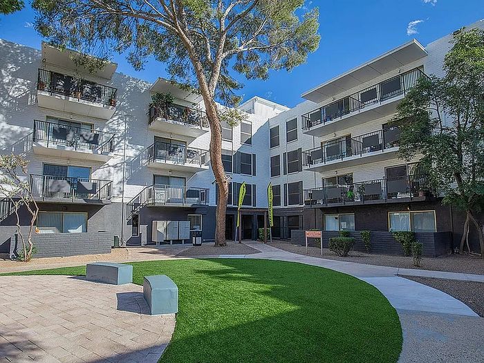 Modern apartment complex with balconies, surrounded by lush greenery and a charming courtyard, under a bright blue sky.