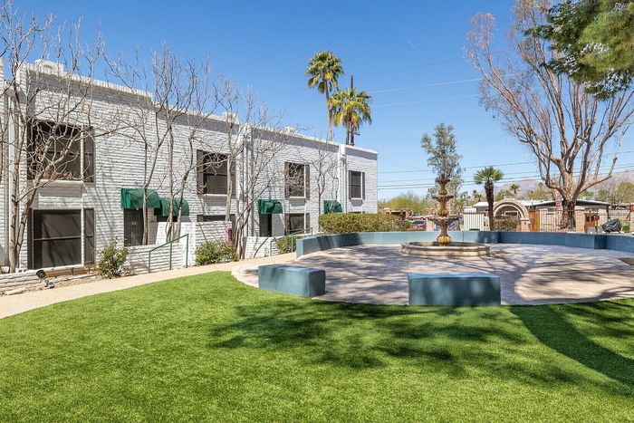 Charming apartment complex with lush green lawn and elegant central fountain, surrounded by well-maintained landscaping and tree-lined paths.