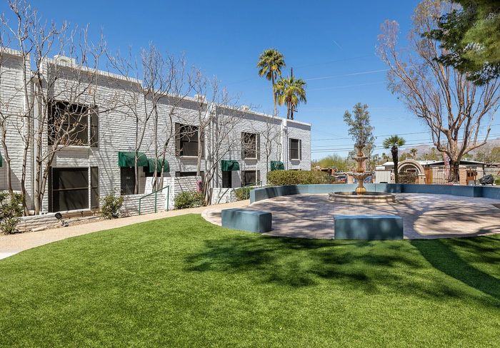 Charming apartment complex exterior with lush green lawns, elegant landscaping, and a central decorative fountain under clear blue skies.