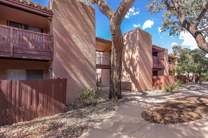 Charming apartment exterior with rustic wooden fencing, shaded by mature trees, featuring private balconies and a sunny pathway.