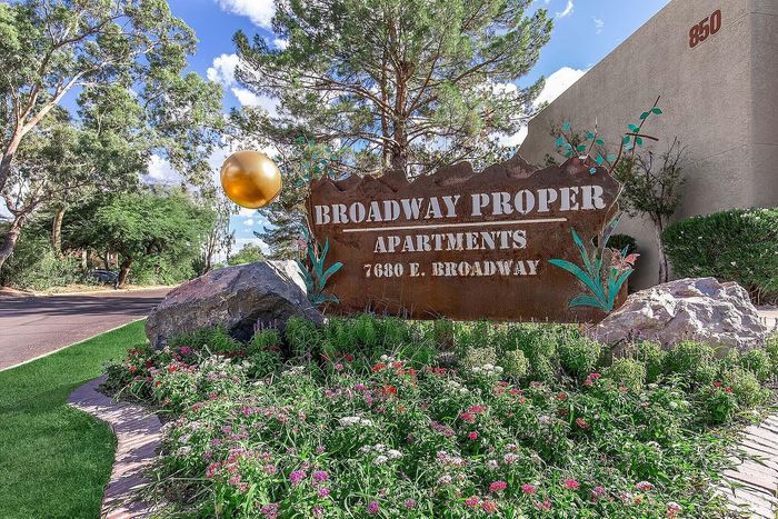 Charming entrance of Broadway Proper Apartments, featuring vibrant landscaping with colorful flowers and elegant signage nestled among lush greenery.