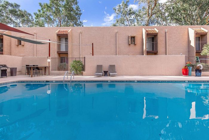 Elegant apartment complex featuring a pristine swimming pool with lounge chairs and outdoor dining area, surrounded by lush greenery and stylish architecture.