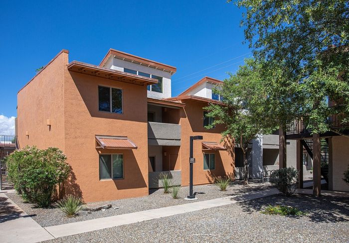 Modern apartment building with a vibrant terracotta exterior, featuring multiple balconies and lush greenery under a clear blue sky.