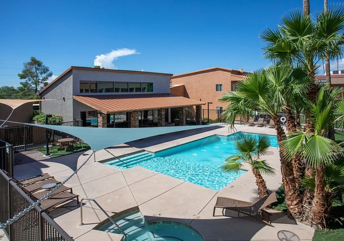 Luxurious apartment complex featuring a sparkling outdoor pool surrounded by lush palm trees, with stylish sun loungers and a shaded patio area.