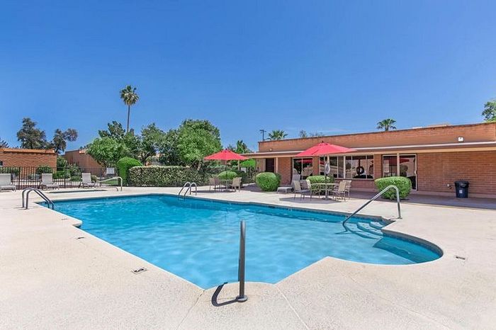 Inviting outdoor swimming pool area with vibrant red umbrellas and comfortable seating, surrounded by lush greenery under a clear blue sky.