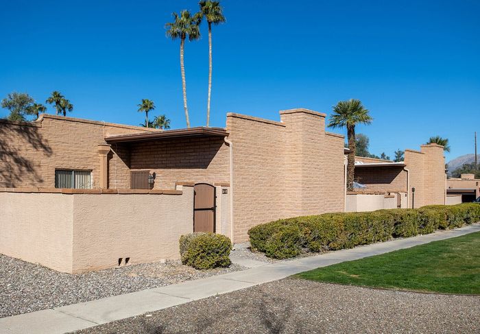 Charming desert townhouse with brick facade, enclosed patio, and lush surrounding greenery under a clear blue sky.