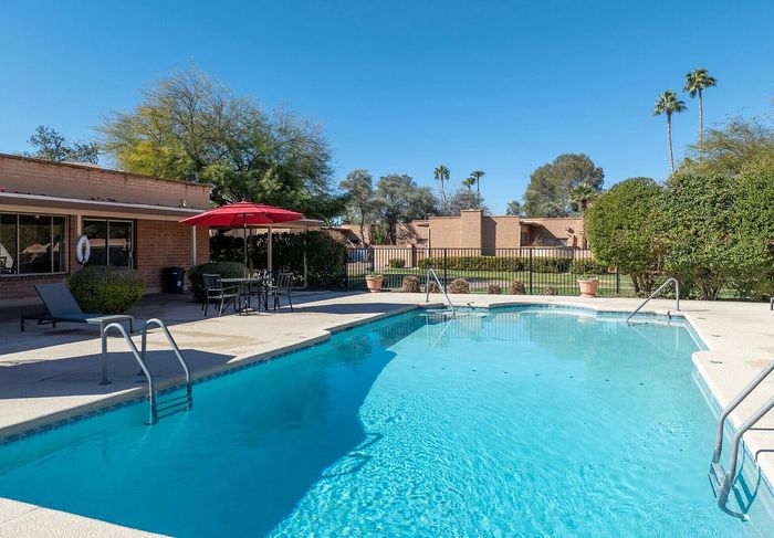 Inviting outdoor pool area with sun loungers, vibrant blue water, and a shaded seating area with umbrella surrounded by lush greenery and clear skies.