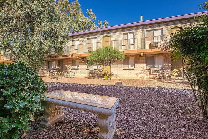 Charming apartment exterior with a serene garden view and inviting stone bench, framed by lush greenery and a warm, sunlit ambiance.