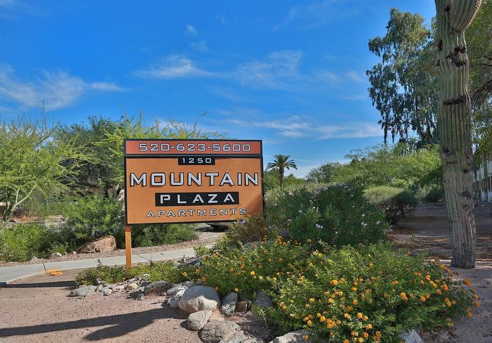 Inviting entrance to Mountain Plaza Apartments, featuring a vibrant desert landscape with lush greenery and colorful flowers under a clear blue sky.
