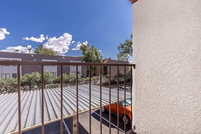 Charming apartment balcony view with blue skies, lush greenery, and a vibrant orange car parked below.