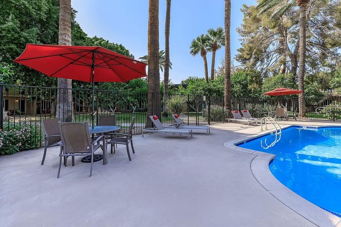 Inviting outdoor pool area with red umbrellas and sun loungers, surrounded by lush greenery and tall palm trees.