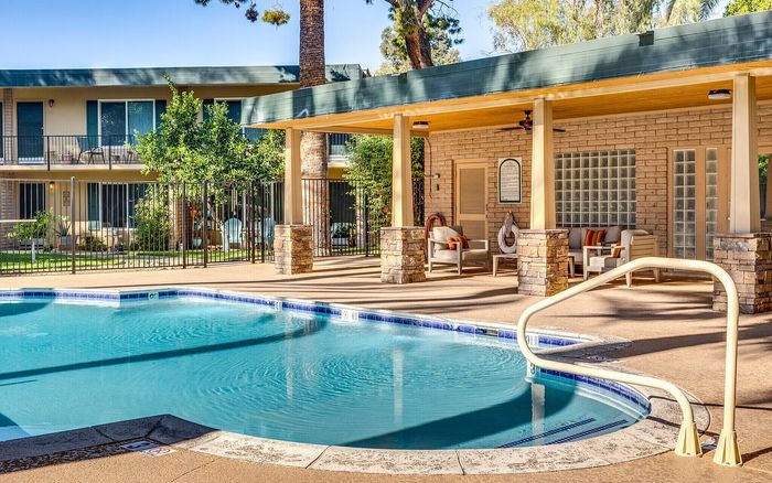 Sunlit apartment complex with elegant poolside seating, surrounded by lush greenery and modern architectural design.