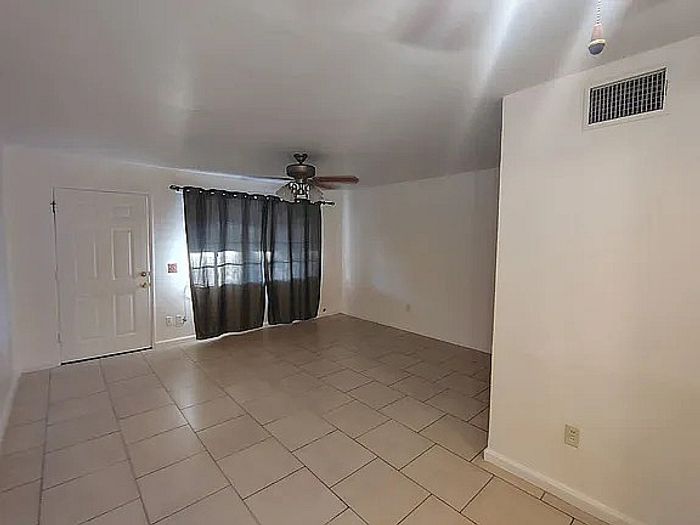 Bright, spacious living room with elegant tiled flooring, ceiling fan, and large window draped with dark curtains for a modern touch.