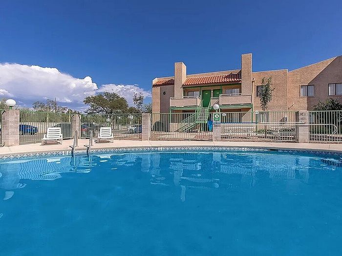 Inviting apartment complex with a sparkling blue swimming pool, sun loungers, and vibrant, green surroundings under a clear sky.