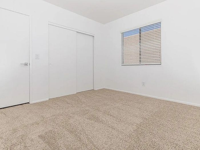 Bright and cozy bedroom with plush carpet, large window with blinds, and sleek white sliding closet doors.