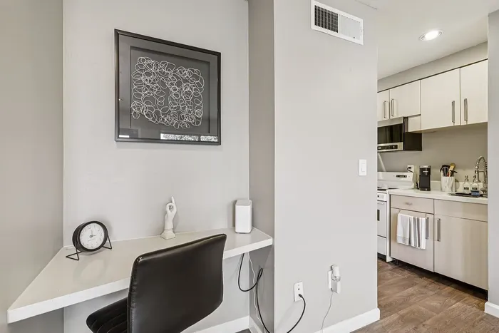Modern apartment corner showcasing a sleek workspace with artistic decor, adjacent to a stylish, fully-equipped kitchen with contemporary cabinetry and warm wooden flooring.