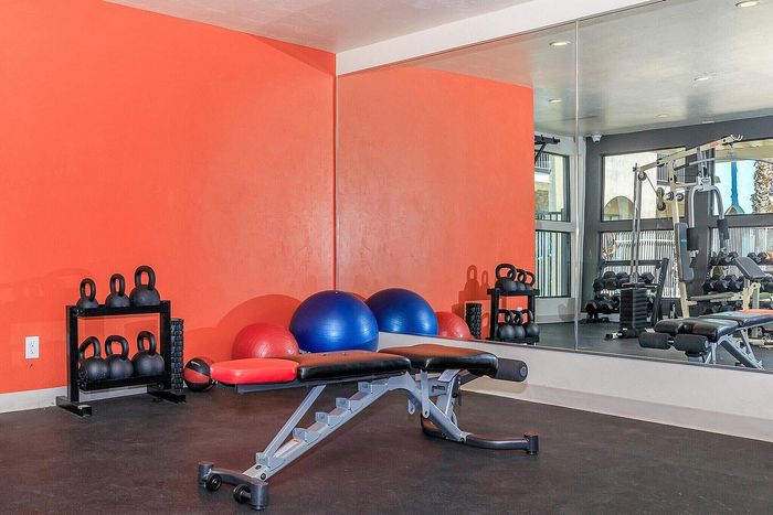 Vibrant home gym with a bold orange accent wall, kettlebells, exercise balls, and a sleek weight bench facing a large mirror.