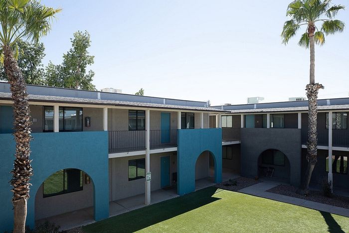 Charming apartment complex courtyard featuring modern architecture with vibrant blue accents, lush green lawn, and tall palm trees under a clear sky.