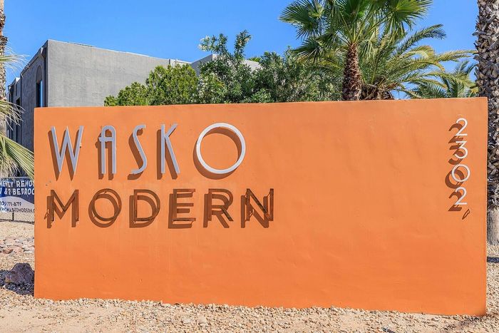 Vibrant orange sign of Wasko Modern apartments framed by lush greenery and palm trees, showcasing a contemporary and stylish living environment.