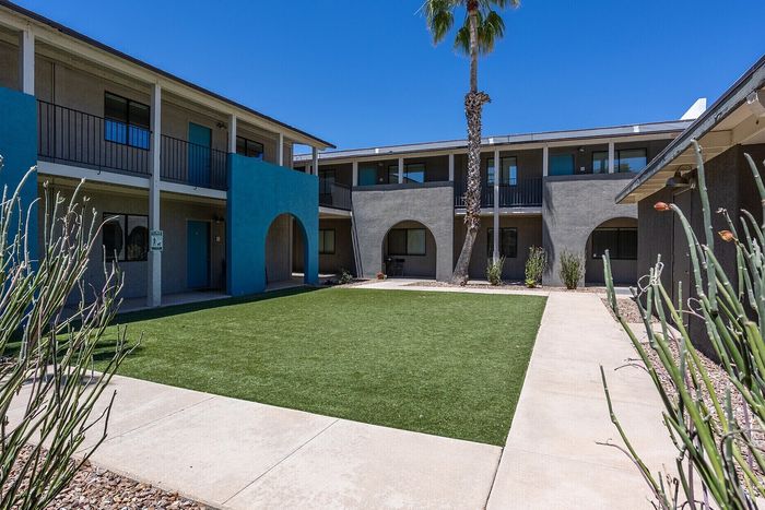Charming apartment complex with modern architecture, vibrant blue accents, and lush green courtyard under a clear blue sky.