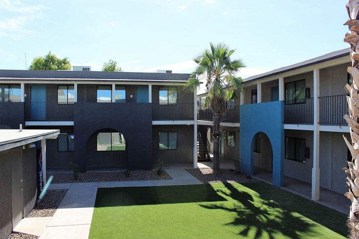 Modern apartment complex with a vibrant courtyard, featuring lush green grass and palm trees under a clear blue sky.