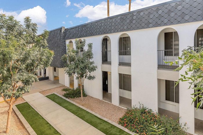 Charming apartment complex exterior with modern design, featuring arched balconies, lush greenery, and a picturesque walkway under a sunny sky.