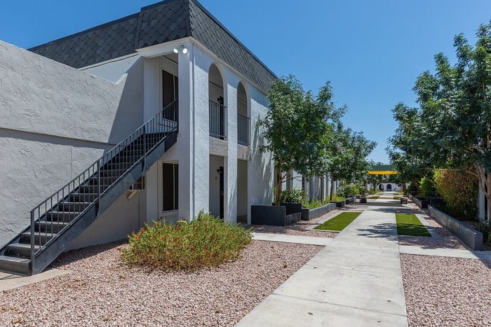 Elegant apartment entrance featuring modern architecture, lush greenery, and a welcoming walkway under a clear blue sky.